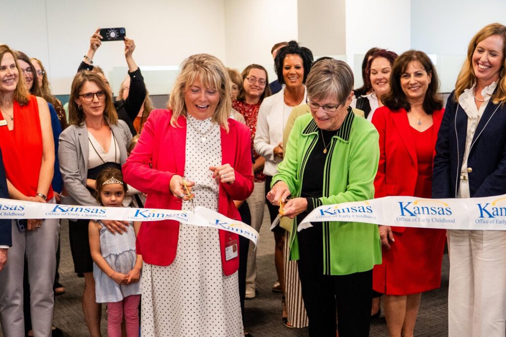 Surrounded by people celebrating the opening of the Office of Early Childhood, Governor Laura Kelly cuts a ceremonial ribbon.